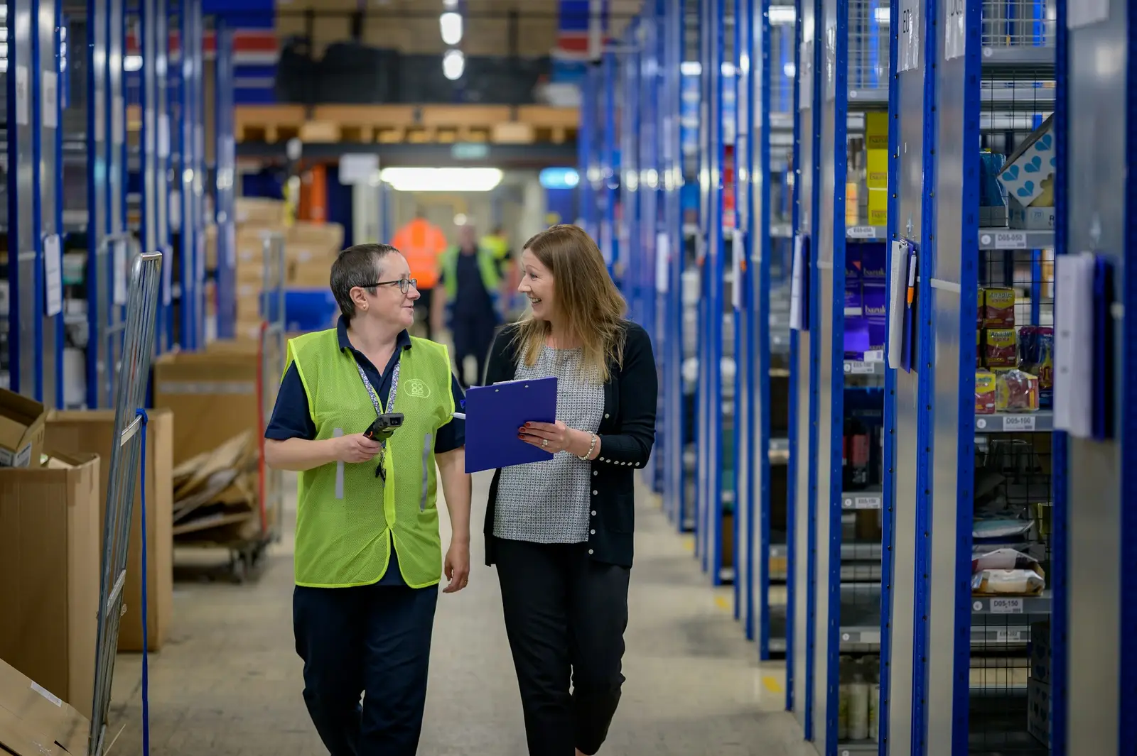 Two warehouse staff walking along an aisle of shelving, one holding a clipboard